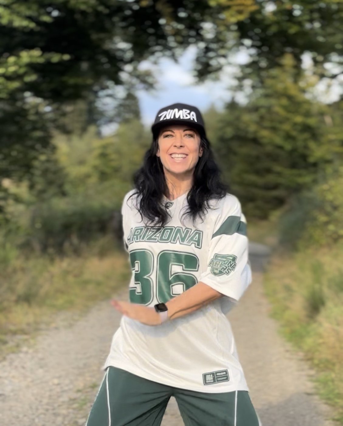 Elena Minich tanzt lachend auf einem Waldweg, Zumba-Cap und weißes Trikot